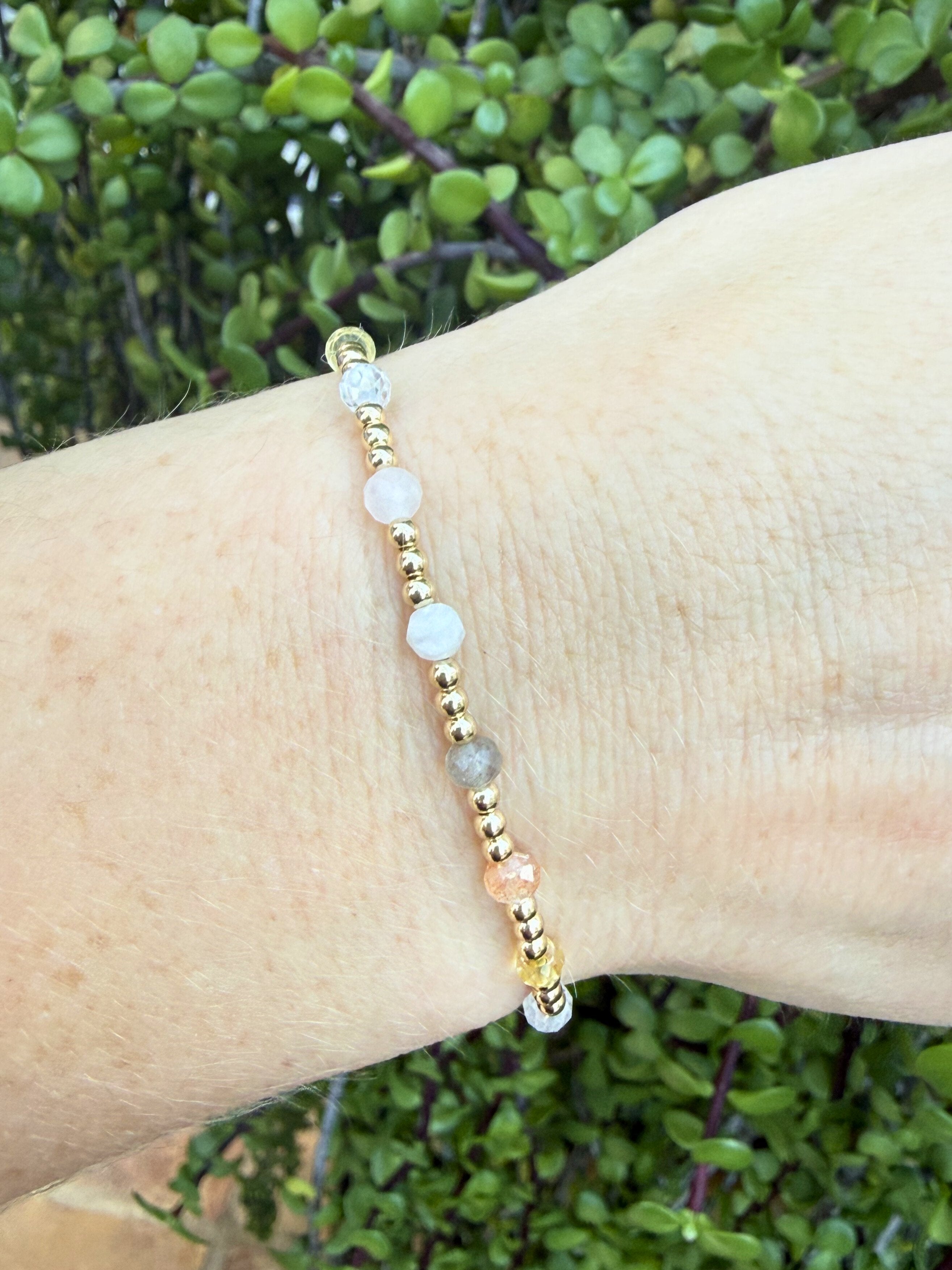 Bracelet with gold, pink, gray, and white beads, a mix of moonstone, sunstone, citrine, zircon, rose quartz, and labradorite on a wrist against a green leafy background