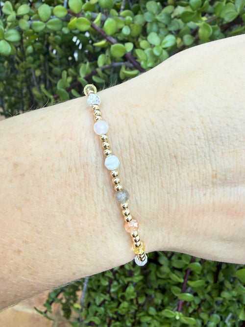Bracelet with gold, pink, gray, and white beads, a mix of moonstone, sunstone, citrine, zircon, rose quartz, and labradorite on a wrist against a green leafy background