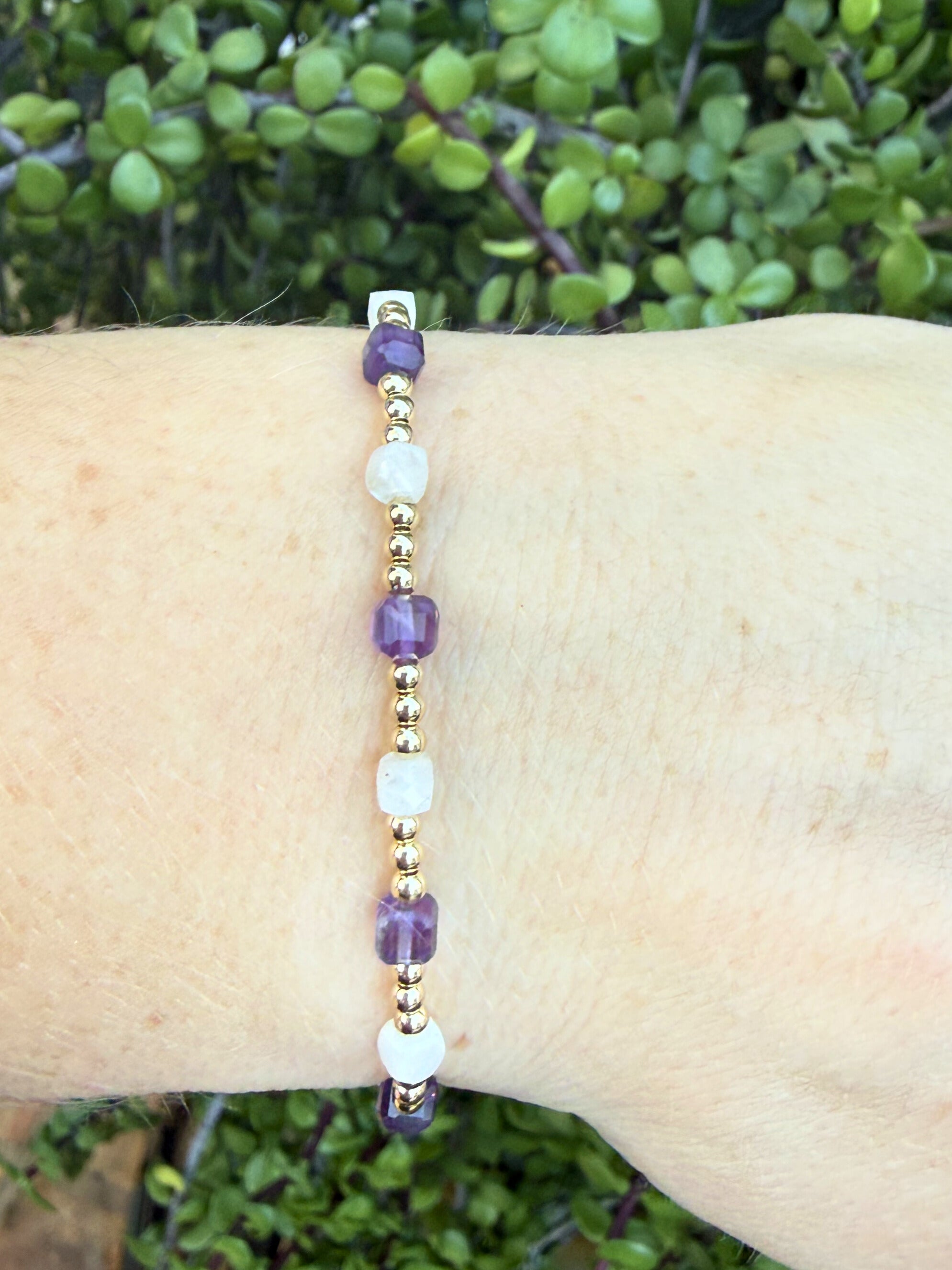 Beaded bracelet with purple and white beads, amethyst and moonstone on a wrist against a green leafy background