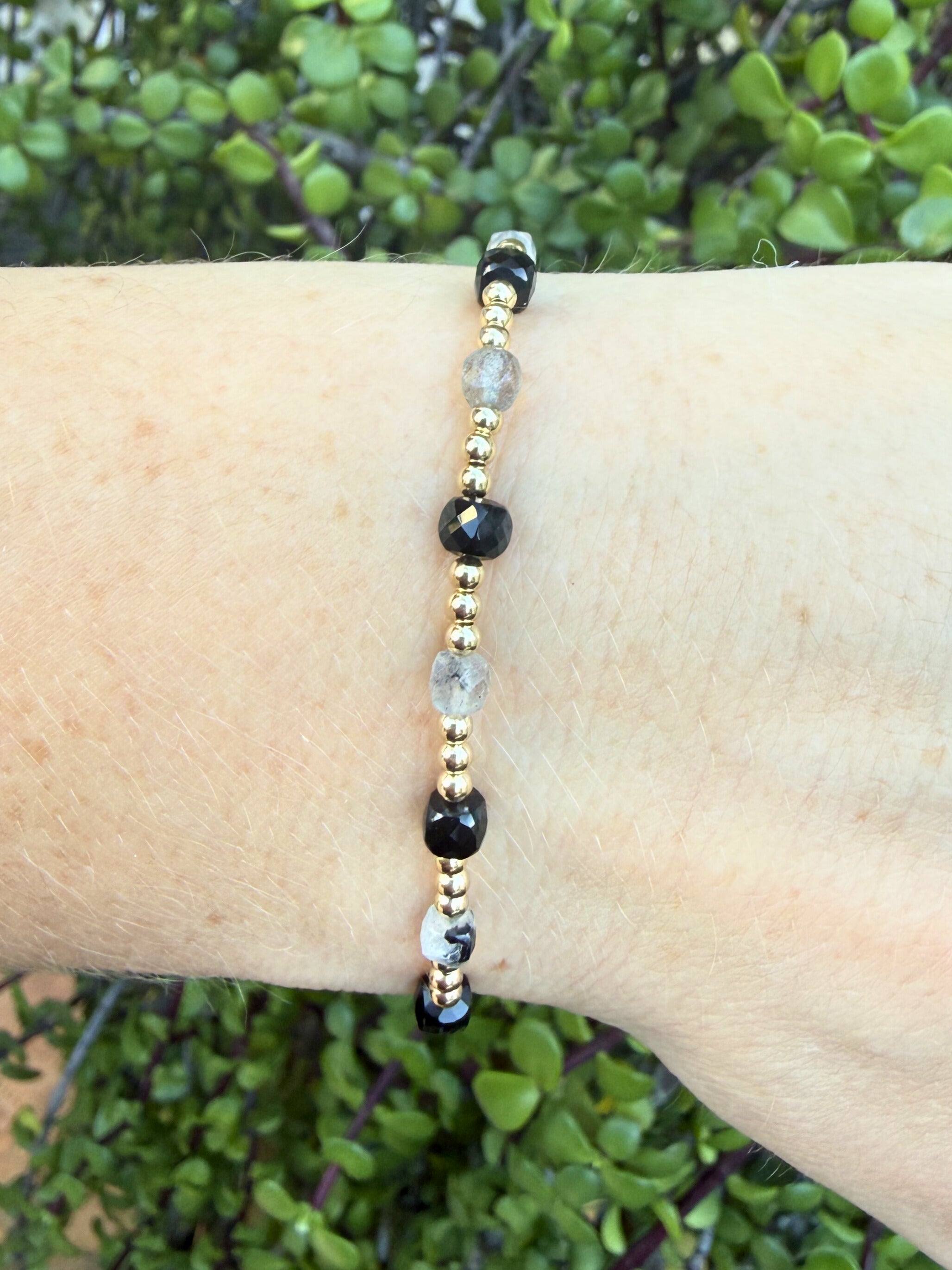 Bracelet with black, gold, and clear beads, black onyx and labradorite on a wrist against a green leafy background