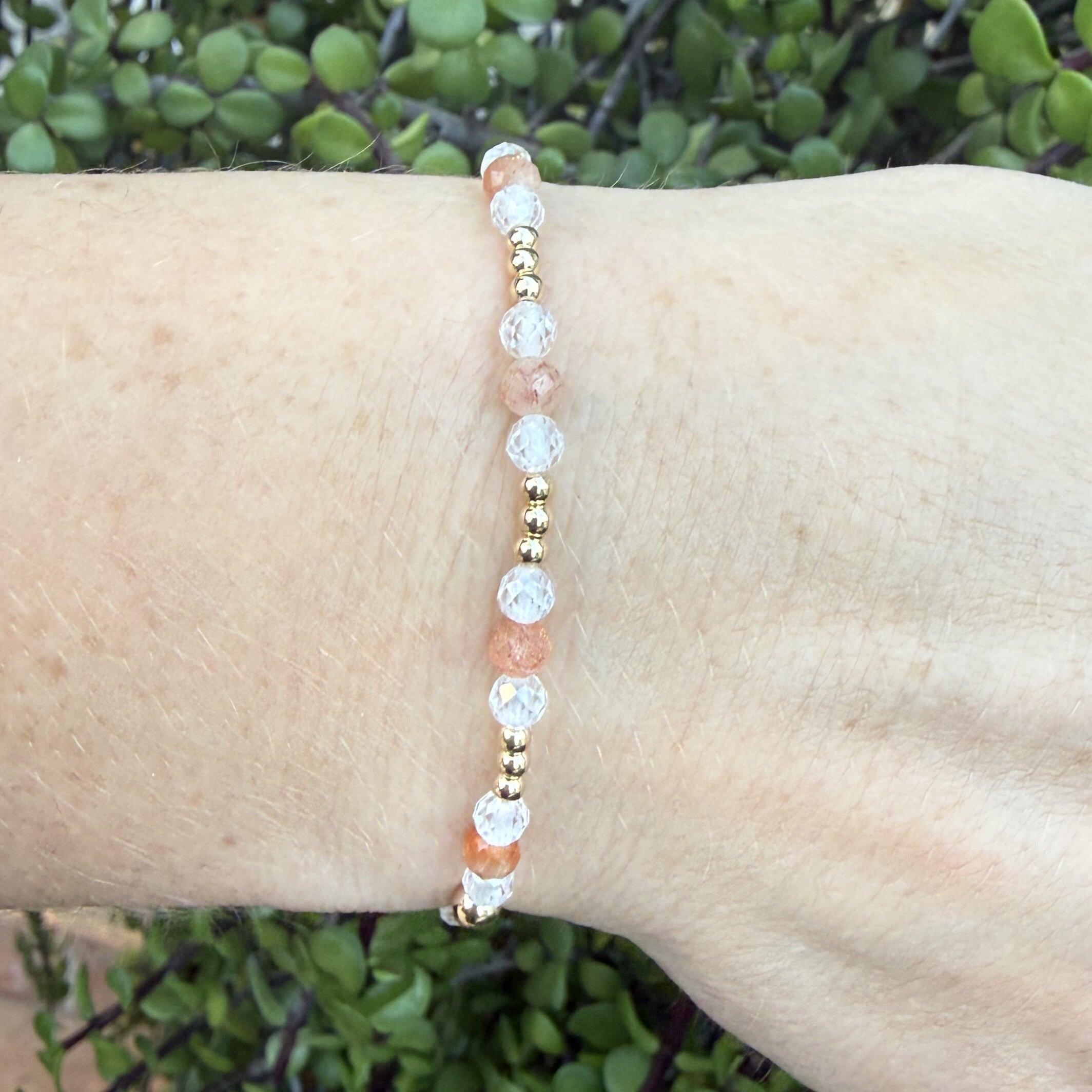Bracelet with peach and gold beads, sunstone and zircon on a wrist against a green leafy background