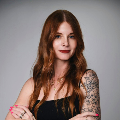 Woman with tattoos and red hair wearing a black top against a gray background